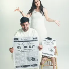 A fun and creative family announcement. While the parents look on, the older sibling hides behind a "Baby Arriving Soon" newspaper prop, adding a playful touch to the photo.