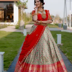 A wide shot of an actress in a stunning red and cream lehenga. The composition uses the pathway to lead the viewer's eye, showcasing the full bridal ensemble in a beautiful outdoor venue.