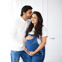 A sweet and casual maternity portrait. The couple's coordinated denim and white outfits create a fresh, modern look against a clean white studio background.