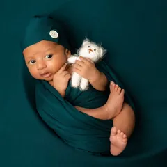 Wide-eyed and wonderful. This awake newborn is nestled in a moon prop, wrapped in a deep teal swaddle and holding a tiny teddy bear, looking right at the camera.
