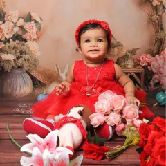 Dressed in a beautiful red dress, this baby girl is the star of her floral-themed sitter session. The studio backdrop is filled with soft flowers and props, creating a sweet and classic portrait that highlights her happy smile.