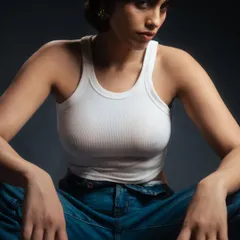A close-up studio portrait with a moody, dark grey background. The lighting is sculpted to emphasize the model's features and the texture of the white tank top, creating an intense and intimate feel.