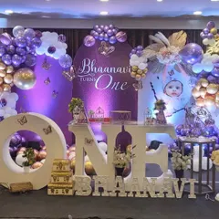 An elegant butterfly-themed first birthday celebration. The decor features a purple and gold color scheme, elaborate balloon arrangements, large "ONE" lettering for photos, and a beautifully decorated dessert table.