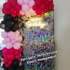 A close-up of a silver shimmer wall with a 'Happy Birthday' neon sign, framed by a pink, fuchsia, and black balloon arch. This creates a high-impact photo corner for any party.