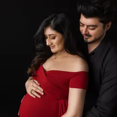 A classic and intimate couple's maternity portrait. Against a simple black background, the father-to-be looks down lovingly at his partner as they both gently hold her belly, highlighted by the striking red of her gown.