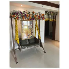 A beautifully decorated swing (jhoola) for a Mundan ceremony, adorned with yellow floral strings and a lush overhead flower arrangement. A portrait of a deity hangs in the background, adding a spiritual touch.