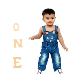 A toddler stands proudly next to the word 'ONE' during his first birthday photoshoot, dressed in cute denim overalls.