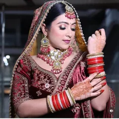 A classic bridal pose in a deep maroon velvet lehenga with intricate gold embroidery. The look is completed with a heavy Polki and ruby choker set, maang tikka, and traditional red bridal bangles.