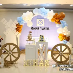 A charming farm-themed birthday party with a barn-shaped backdrop and a color scheme of light blue, white, and brown. The decor includes cutouts of a cow and sheep, large wagon wheels, and illuminated letters on a bed of hay.