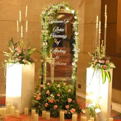 A personalized welcome for an intimate Roka. A floral-framed mirror with the couple's names is flanked by illuminated pedestals and candles, creating a warm and inviting entrance.