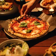 A hand reaches for a slice of freshly baked Margherita pizza, the centerpiece of a warm, shared meal. Our private dinners are about heartfelt flavors and creating moments of connection.
