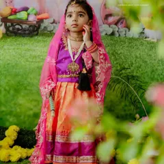 This little Radha looks thoughtful and divine in her colorful outfit. The soft, dreamy framing through the leaves adds an artistic touch to this festive portrait.
