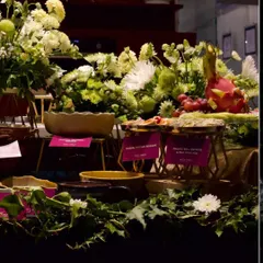An evening grazing table setup, where lush green and white floral arrangements complement the rich colors of the food. You can see labeled dishes like "Smoked Baba Ganoush" and "Dragon Fruit, Melon & Feta Salad," showcasing the diverse menu.
