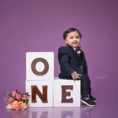 Celebrating a first birthday milestone with a classic studio portrait. A smartly dressed one-year-old boy sits proudly on large "ONE" blocks against a clean purple background, creating a timeless memory of this special day.