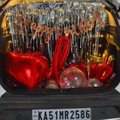A romantic car boot decoration to surprise a wife on her birthday. This setup features red heart-shaped foil balloons, a "Happy Birthday" banner, and fairy lights against a silver shimmer curtain.