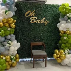 An elegant "Oh Baby" shower setup with a lush green foliage wall. The backdrop is framed by beautiful balloon columns in shades of green, gold, grey, and white, creating a fresh and natural look.