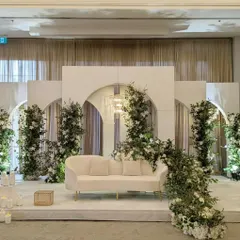 A minimalist and elegant stage design for a modern reception or Nikkah ceremony. I used clean white panels with archways, accented with fresh greenery and soft lighting to create a sophisticated and serene ambiance.