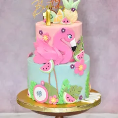 A tropical two-tier cake for a flamingo-themed first birthday. This vibrant creation is adorned with a hand-sculpted pink flamingo, fondant pineapples, watermelons, and tropical leaves for a fun, summery celebration.