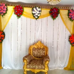 A simple flower decoration for a Half Saree ceremony. The backdrop of yellow and white drapes is accented with colorful flower bouquets and a traditional gold-colored chair.