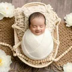 A smiling newborn swaddled in white, nestled in a basket with boho-style macrame and floral details. This setup is all about natural textures and pure sweetness.