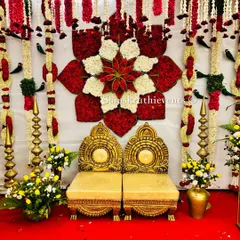A close-up of a wedding mandap backdrop featuring a large, intricate floral lotus made from red and white petals. The traditional golden chairs are placed for the bride and groom, completing the sacred look.