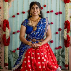 A bride in a vibrant half-saree for a traditional ceremony. The backdrop features intricate floral work that complements her attire perfectly.