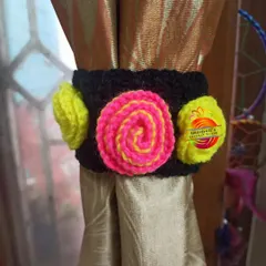 A modern and vibrant curtain tie-back design. It features a black band decorated with a central pink and yellow swirl and two bright yellow pom-poms on the sides.