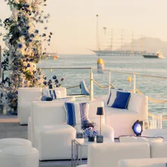 The lounge setup for the welcome dinner, with comfortable white sofas, blue and white striped cushions, and a floral arch, all set against the golden hour glow on the water.
