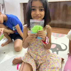 A young girl proudly holds up the colorful, textured material she created in a workshop. The pride in her creation is evident.