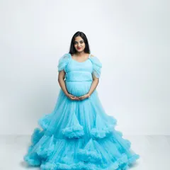 A beautiful solo maternity portrait featuring a stunning, layered blue gown. The clean white studio background makes the color pop, creating a bright, airy, and elegant photograph that celebrates the mother-to-be.