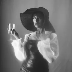 A dramatic, high-fashion monochrome portrait. The model wears a large, floppy hat and holds a glass of wine, creating a look of classic, old-Hollywood glamour with a modern, moody twist.