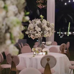 As evening falls, the same reception setup is transformed by soft lighting. The large white floral centerpieces seem to glow in the twilight, creating a romantic and intimate atmosphere for the dinner celebration.