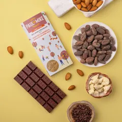 A bright and clean shot of an almond chocolate bar on a yellow background, with raw ingredients like cocoa beans and almonds.