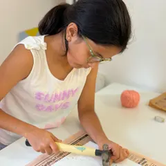 A young artist learns to use a hammer safely to place the nails for her initial string art project, developing new skills and concentration.