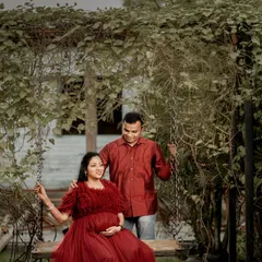 A romantic outdoor shot of the couple on a swing surrounded by lush greenery. The mom-to-be's flowing red gown adds a beautiful pop of color to the natural scene.