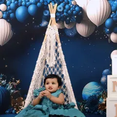 A baby girl sits in a macrame swing for her first birthday, surrounded by a stunning blue and white balloon arrangement and 'ONE' blocks.