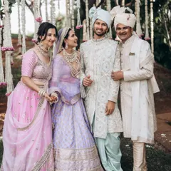 A bride and groom pose with their parents under a beautiful floral arrangement. The coordinated pastel outfits and genuine smiles make this a perfect multi-generational family portrait from a wedding day.