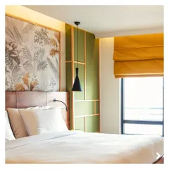 The bedroom at the Green Bean Hotel features a custom wallpaper with a tropical leaf motif, paired with a wooden headboard and green paneled wall. The design aims to create a serene and restful sanctuary for guests.