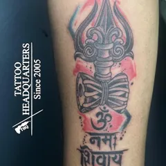 A modern take on a spiritual tattoo, this design features a Trishul and Damaru with "Om Namah Shivaya" in Devanagari script, accented with a splash of color.