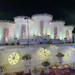 An elegant, all-white wedding stage design that feels fresh and sophisticated. The backdrop uses flowing white drapes and panels of pink and white flowers, with unique illuminated floral lights in the foreground.