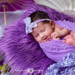 A precious newborn girl gives a little smile while sleeping in a purple-themed setup. She is nestled in a large white shell prop, surrounded by purple fur and flowers.