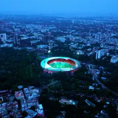 An aerial view of the M. Chinnaswamy Stadium at dusk, the heart of cricket in Bengaluru. This shot captures the stadium nestled within the city's green canopy, lit up and ready for action, showcasing its iconic status.