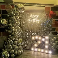 A stylish 21st birthday setup featuring a silver shimmer wall, a "Happy Birthday" neon sign, and a chrome silver balloon garland. The large light-up marquee numbers make a bold statement.