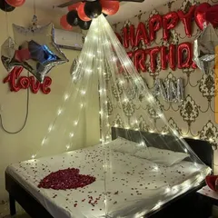 A classic romantic birthday setup featuring a canopy with fairy lights over the bed, which is decorated with a heart made of rose petals. The red and black balloons, along with "Happy Birthday" and "Love" foil letters, make the message clear.