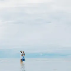 A minimalist pre-wedding photo featuring the couple standing in calm, shallow water. The vast, overcast sky and serene sea create a peaceful and romantic atmosphere.