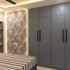 A master bedroom in Bhayandar West with a cool grey color palette. The room includes a large four-door wardrobe, a textured wallpaper accent wall with backlighting, and open shelves for displaying personal items.