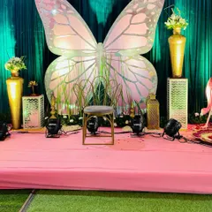A whimsical stage setup for a baby shower, featuring a giant pink and iridescent butterfly as the main backdrop. The stage is decorated with golden vases and floral arrangements.