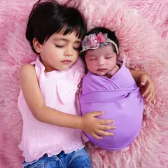 A heart-melting moment of pure love as an older sister sleeps beside her swaddled newborn sibling on a soft pink blanket.