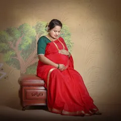 A serene portrait of a mother-to-be in a classic red saree. This studio shot uses a painted backdrop and soft lighting to create a warm, traditional feel.