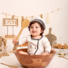 A happy chef in his kitchen. This little one holds up a wooden spoon with a big smile, surrounded by baked goods and kitchen props in this delightful themed shoot.